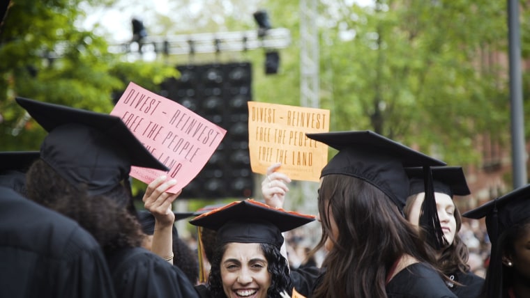 In a show of protest, Harvard graduates hold up red and orange signs that say, "Divest - reinvest free the people free the land" while university president Larry Bacow speaks.
